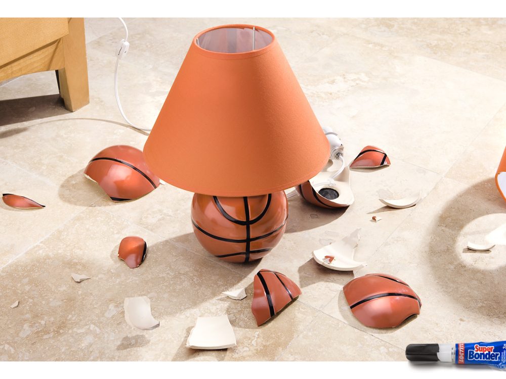 Una lámpara nocturna infantil con forma de balón de basquetball intacta. Alrededor vemos pedazos de otra lámpara igual. Abajo vemos un pegamento.